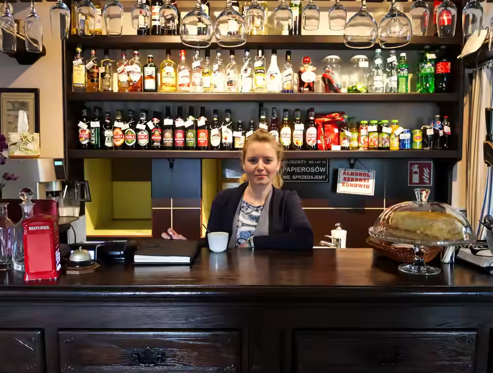 Bar w Karioce: lada z butelkami na półkach i szklankami zawieszonymi nad barem, za ladą uśmiechnięta barmanka